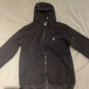 Svart hoodie från Polo Ralph Lauren S - Svart hoodie från Polo Ralph Lauren med dragkedja och huva. Klassisk vit logga broderad på bröstet, två fickor framtill och snörning i huvan. Perfekt basic-plagg med clean look och bekväm passform.