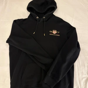Svart hoodie från GANT - En mycket fin hoodie från Gant i nästintill nyskick. Relativt lång i ärmarna så perfekt för de lite längre. Axelbredd: 54cm Längd: 71 cm