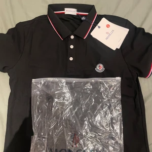 Svart Moncler pikétröja med röd/vit detalj - Svart pikétröja från Moncler i bomull med klassisk krage och knäppning framtill. Röd och vit rand på krage och ärmslut, samt Moncler-logga på bröstet. Perfekt för dig som gillar stilrena och exklusiva plagg.