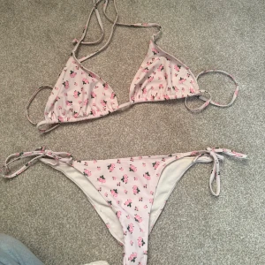 Ljusrosa blommig bikini med knyt - Supersöt ljusrosa bikini från Nelly med små rosa blommor och gröna blad. Triangeltopp med smala axelband och knyt i ryggen, samt matchande bikinitrosa med knyt i sidorna. Perfekt för stranden eller poolen! Storlek S i underdelen och M i överdelen!