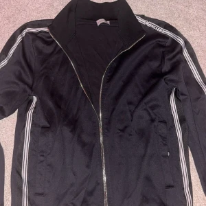 Svart Moncler trackjacka med ränder - Svart trackjacka från Moncler med vita ränder längs ärmarna och dragkedja framtill. Jackan har hög krage, Moncler-logga på axeln och är tillverkad i bomull och polyester. Perfekt för en sportig och clean look. Limited kan även byta mot något går ner i pris vid snabb affär