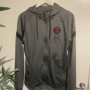 PSG grå vindjacka med huva - Grå vindjacka från PSG med svart och rött klubbmärke på bröstet, hel dragkedja och huva med snörning. Jackan har svarta detaljer på ärmarna och praktiska fickor med dragkedja. Perfekt för dig som gillar sportig stil och vill representera Paris Saint-Germain.