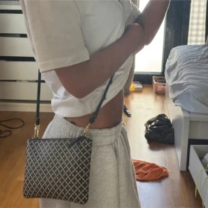 Svartvit taske fra Malene Birger  - Säljer en stilren axelväska med svartvitt rutigt mönster och guldfärgade detaljer. Väskan har justerbar axelrem i svart och stängs med dragkedja. Perfekt storlek för mobilen och det viktigaste. Materialet känns som syntet med struktur.