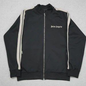 Palm Angels svart track jacket - Svart track jacket från Palm Angels med vita ränder längs ärmarna och logga på bröstet. Jackan har hel dragkedja, ribbade muddar och två fickor med dragkedja. Perfekt för en sportig och trendig streetstyle-look.