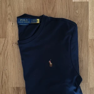 Mörkblå t-shirt från Polo Ralph Lauren - Snygg mörkblå t-shirt från Polo Ralph Lauren i bomull med klassisk broderad polospelare på bröstet. Modellen är custom slim fit och har rund halsringning och korta ärmar. Perfekt för en clean och stilren look.
