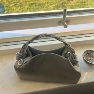 Grå axelväska med flätat handtag - Snygg grå axelväska i syntetmaterial med unikt flätat handtag och silvriga metalldetaljer. Väldigt lik Ganni väska