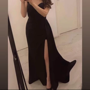 Svart långklänning med slits - Elegant svart långklänning med axelbandslös design och hög slits framtill. Klänningen har en figurnära passform upptill och faller snyggt längs kroppen. Perfekt för dig som vill ha en stilren och sofistikerad look. Den är använd en gång!