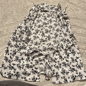 Blommig väst med volang från SHEIN - Snygg vit väst med svarta blommor från SHEIN. Västen har volangdetaljer längs axlarna och knytning framtill. Perfekt för att ge outfiten en romantisk touch. Lätt och luftig känsla, passar till många olika looks.