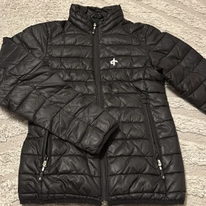 Svart quiltad dunjacka från Cross - Svart dunjacka från Cross med quiltad design och hög krage. Jackan har två dragkedjeförsedda fickor och en diskret logga på bröstet. Perfekt för kyliga dagar och enkel att matcha med olika outfits.