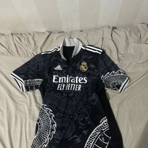 Real Madrid svart draktröja Adidas - Snygg svart Real Madrid fotbollströja från Adidas med coolt drakmönster i grått. Tröjan har korta ärmar, vit krage och detaljer samt klubbmärke i guld på bröstet. Materialet är lätt och andas, perfekt för match eller träning. Är is storlek L. Prisk kan diskuteras!