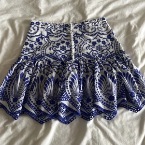 Vit och blå mönstrad kjol från Zara - Supersöt kortkjol från Zara i vitt och blått med unikt mönster och volangig nederdel. Kjolen har dekorativa knappar framtill och är gjord i bomull, perfekt för dig som gillar statement-plagg med snygga detaljer.