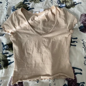 Beige v-ringad topp med volangkant - Säljer en beige topp med v-ringning och korta ärmar. Toppen har volangkant vid ärmslut och nederkant, vilket ger en söt och trendig look. Perfekt att matcha med jeans eller kjol för en chill stil.
