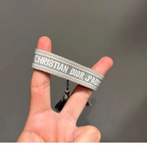 Grått armband Christian Dior J'Adior - Snyggt vävt armband från Christian Dior i ljusgrått med texten 'J'Adior' broderad i vitt. Armbandet har justerbar knytning med tofsar i ändarna för en cool och trendig look.