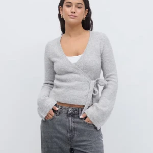 Grå omlottkofta med knyt - Supermjuk grå omlottkofta med knyt i sidan och v-ringad hals. Koftan har långa ärmar med lite vidare avslut och är stickad i ett fluffigt material. Perfekt att slänga på sig över ett linne eller t-shirt för en chill look.