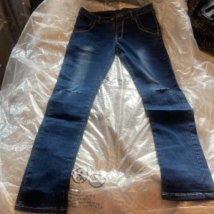 Blå jeans Ancient Dragon stl 30 - Mörkblå jeans från Ancient Dragon med snygga slitningar vid knäna och kontrastsömmar. Klassisk femficksmodell med rak passform och läderpatch bak i midjan. Jeansen är tillverkade i ett stretchigt bomullsmaterial för skön känsla.