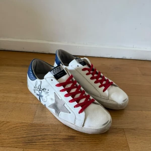 Golden Goose vita sneakers med röd snörning - Golden Goose sneakers i vitt skinn med grå stjärndetalj på sidan, blå hälkappa och röda skosnören. Sidan har svart grafittiliknande print och sulan är platt. Märket syns på häl och plös. Perfekta för dig som gillar streetstyle med en twist.
