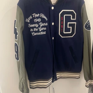 Gant bomberjacka Anniversary Edition - Snygg bomberjacka från Gant Anniversary Edition med mörkblå kropp och olivgröna ärmar. Stora broderade detaljer, randiga muddar och knappar framtill. Stor G-logga på bröstet och broderad text på både utsida och insida. Perfekt för dig som gillar sportig och retro stil.