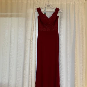 Vinröd långklänning med spetsdetaljer - Supersnygg vinröd långklänning med v-ringad spetsliv och blommiga broderier. Klänningen har breda axelband och figurnära passform upptill. Perfekt för bal eller fest, med eleganta detaljer och romantisk känsla.