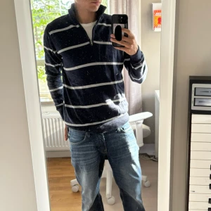 Randig marinblå half zip tröja - Säljer en marinblå tröja med vita horisontella ränder och half zip i halsen. Tröjan har hög krage, ribbade muddar och är långärmad. Perfekt för dig som gillar en avslappnad men ändå stilren look.