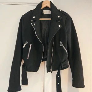 Svart mocka jacka från NLY Trend - Snygg svart jacka i mocka från NLY Trend med silvriga dragkedjor, bälte i midjan och coola detaljer. Jackan har lång ärm och klassisk krage med tryckknappar. Perfekt för dig som vill ha en edgy look.