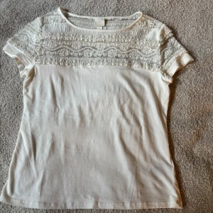 Vit blus med spets från H&M - Säljer en söt vit blus från H&M i storlek M. Toppen har korta ärmar och en snygg spetsdetalj🌼🌼