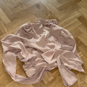 Beige omlottopp från Gina Tricot - Supersnygg beige omlottopp från Gina Tricot i satinliknande material. Toppen har långa puffärmar, hög krage med rynkade detaljer och knytband i midjan. Perfekt för dig som gillar trendiga och feminina plagg med det lilla extra.