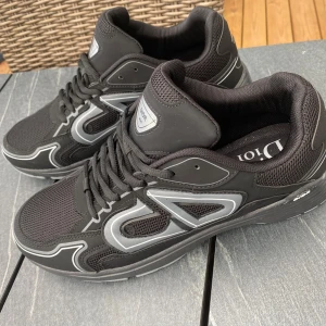 Svarta Dior B30 sneakers med silverdetaljer - Dior B30 sneakers i svart med silvriga detaljer och tydlig Dior-logga på sidan. Skorna har meshpaneler för extra andningsförmåga, snörning och en robust, grå sula. Perfekta för dig som gillar exklusiva och sportiga sneakers med modern design.