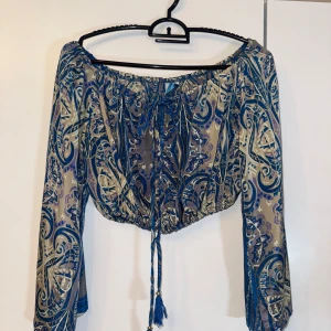 offshoulder blus - Supersnygg offshoulder blus från Ble med blått och beige paisleymönster. Croppad modell med långa vida ärmar och knytning framtill. Tunn och luftig, perfekt för varma dagar eller festival. Designad i Grekland och ger en riktigt bohemisk vibe. Har inga defekter överhuvudtaget. Pris kan diskuteras 
