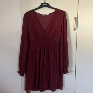 Vinröd omlottklänning från NA-KD - Supersnygg vinröd kortklänning från NA-KD med omlott och v-ringning. Klänningen har långa ärmar, smockad midja och lätt volangdetalj. Tunt, flowy tyg som ger en luftig känsla. Perfekt för dig som vill ha en chic och trendig look.