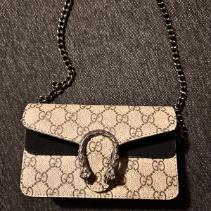 Gucci Dionysus axelväska med kedja - Säljer en Gucci Dionysus axelväska med ikoniskt GG-mönster i beige och brunt. Väskan har detaljer i svart skinn, silverfärgad kedjerem och ett snyggt spänne framtill med ormhovedesign. Perfekt storlek för det viktigaste och riktigt lyxig känsla.