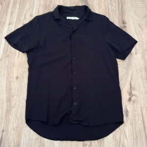 Svart kortärmad skjorta med knappar - Stilren svart skjorta från ”the Blue uniform”  med korta ärmar och klassisk krage. Skjortan har knäppning framtill och är tillverkad i ett mjukt, lätt material (viskos)
