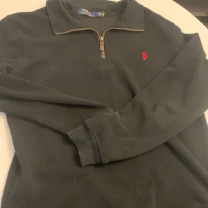 Svart half zip tröja från Polo Ralph Lauren - Svart långärmad tröja från Polo Ralph Lauren med röd broderad logga på bröstet. Tröjan har en snygg half zip-dragkedja och är gjord i mjukt material som passar perfekt till lager-på-lager. Klassisk och stilren design.