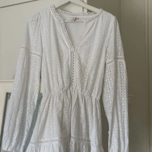 Vit kort klänning med broderade detaljer - Superfin vit kort klänning med broderade hål och volangkant nertill. Klänningen har långärmade puffärmar, markerad midja och v-ringning med spetsdetaljer. Perfekt för sommarens alla tillfällen och student.