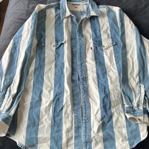 Oversized randig jeansskjorta Levi's - Säljer en oversized jeansskjorta från Levi's med breda blå och vita ränder. Skjortan har två bröstfickor med knappar och klassisk krage. Tillverkad i bomull och har en avslappnad passform som ger en cool och trendig look.