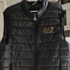 Svart dunväst från Emporio Armani - Snygg svart dunväst från Emporio Armani EA7 med guldfärgad logga på bröstet. Västen är quiltad och har dragkedja framtill. Perfekt att slänga över en hoodie för en cool och sportig look.