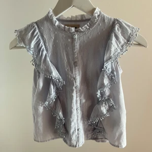 Blå Zadig & Voltaire blus - Supersöt ljus blå blus från Zadig & Voltaire med volanger längs axlarna och fina spetsdetaljer. Blusen har knappar framtill med rysch och diskret strukturmönster i tyget. Perfekt för dig som gillar somrig och trendig stil. Ny pris är 1300 kr.