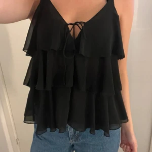 Svart volangig linnetopp med knyt - Supersöt svart linnetopp med flera lager volanger och smala axelband. Toppen har en snygg v-ringning med knyt framtill som ger en extra detalj. Perfekt att styla med jeans eller kjol för en trendig look. Är lite sönder därav priset