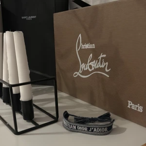 Christian Dior J'Adior armband - Snyggt armband från Christian Dior med texten 'J'Adior' broderat i vitt på en mörkblå bakgrund. Armbandet har justerbar knytning med tofsar i ändarna och är gjort i textil. Perfekt accessoar för att lyfta din outfit.