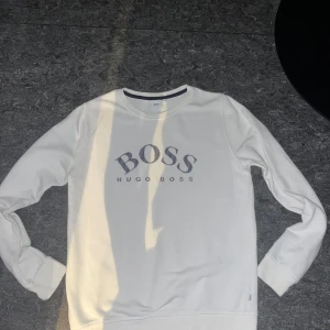 Vit Hugo Boss Sweatshirt - Säljer en stilren vit sweatshirt från Hugo Boss med klassisk rund hals och tryckt logga på bröstet. Tröjan har långa ärmar och ribbade muddar. Perfekt för dig som gillar enkel och snygg design med premiumkänsla.