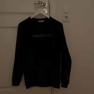 Svart långärmad Moncler tröja - Svart långärmad tröja från Moncler med klassisk logga framtill och diskret röd detalj på ärmen. Tröjan har rund halsringning och är tillverkad i mjukt material som känns skönt mot huden. Perfekt för dig som gillar stilrena och exklusiva plagg.