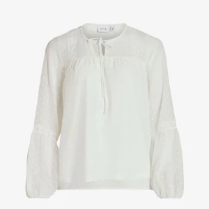 Vit blus med knyt från Vila - Supersöt vit blus från Vila med knyt i halsen och diskreta prickar i tyget. Blusen har långa, luftiga ärmar med spetsdetaljer och lätt transparent känsla. Perfekt för dig som gillar romantisk och bohemisk stil.