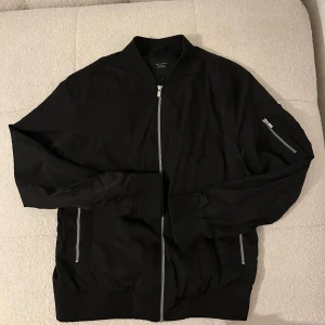 Svart bomberjacka Jack & Jones L - Snygg svart bomberjacka från Jack & Jones Premium i storlek L. Jackan har klassisk ribbad krage, muddar och silverfärgade dragkedjor på framfickor och ärm. Perfekt för dig som gillar stilren och enkel design.