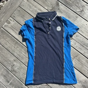 Blå och marinblå polo från Tommy Hilfiger - Snygg kortärmad pikétröja från Tommy Hilfiger i marinblått och blått med vita detaljer. Klassisk krage med ränder, knappar framtill och broderad logga på bröstet. Tröjan är gjord i mjuk bomull och har en slim passform som sitter schysst.