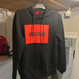 Svart hoodie från HUGO med röd logga - Svart hoodie från HUGO med stor röd logga framtill. Klassisk huva med snörning och känguruficka. Tröjan har lång ärm och en relaxed passform. 