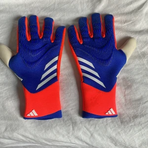 Adidas Predator målvaktshandskar blå/röd - Säljer ett par Adidas Predator målvaktshandskar i blått och rött med vita detaljer. Handskarna har en tight passform, greppvänlig insida och klassisk Adidas-design med tre ränder. Perfekta för fotbollsmålvakter som vill ha stil och funktion på planen. Priset är inte hugget i stenen. 