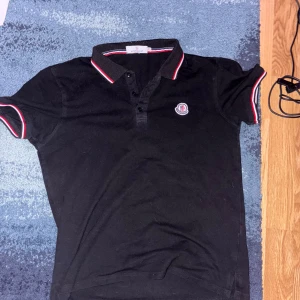 Svart Moncler t-shirt med röd/vit kant - Svart t-shirt från Moncler med korta ärmar och klassisk polokrage. Röd och vit randig kant på krage och ärmslut ger en clean och sportig vibe. Moncler-logga broderad på bröstet. Tillverkad i mjuk bomull för skön komfort.