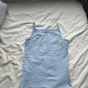 Ljusblått linne från H&M - Supersöt ljusblå linnetopp från H&M med smala axelband och liten rosett framtill. Toppen har en enkel och clean look med diskret spetskant längs ringningen. Perfekt för varma dagar eller som lager under en skjorta.