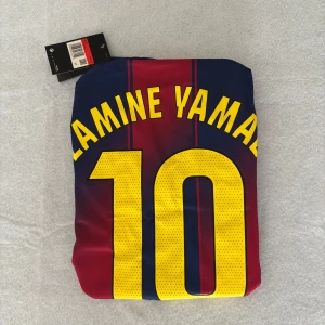 Barcelona Lamine Yamal 10 Nike fotbollströja - Säljer en officiell Barcelona fotbollströja med Lamine Yamal och nummer 10 på ryggen. Tröjan är blå och röd med gula detaljer, tillverkad i Nike Dri-FIT Breathe-material som andas och håller dig torr. Klassisk randig design och perfekt för alla Barca-fans. Känns mer som en M