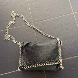 Svart tiamo axelväska med kedja - Cool svart axelväska i skinn med grov silverfärgad kedja som axelrem och detaljer runt kanterna. Väskan har ett fack med dragkedja på baksidan och snygga sömmar längs kanten. Perfekt för dig som gillar en edgy stil.