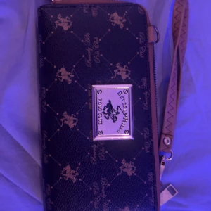 Svart plånbok Beverly Hills Polo Club - Snygg svart plånbok från Beverly Hills Polo Club med guldigt mönster och logga framtill. Plånboken har dragkedja runtom, flera fack inuti och en avtagbar handledsrem i brunt med dekorativ söm. Perfekt för att hålla koll på kort och cash. och har bara använt en gång 💓